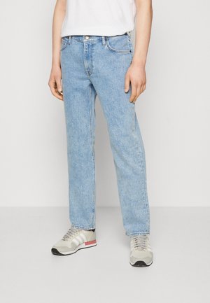 Personne portant un jean droit bleu clair, des baskets blanches avec des accents beige et rose, et un t-shirt blanc, debout devant un fond uni.