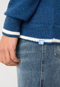 Blå strikket genser med hvit ribbestrikket kant og ermer, med en liten blå etikett nederst. Bært med lyseblå denimjeans.