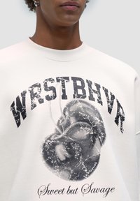 Felpa color crema con una grafica nera audace recante la scritta "WRSTBHVR" e un design centrale che rappresenta un volto. La texture è liscia.