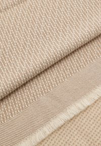 Beige vævet stof med et struktureret mønster, der indeholder horisontale og vertikale linjer. Kanterne er minimale med en blød finish.