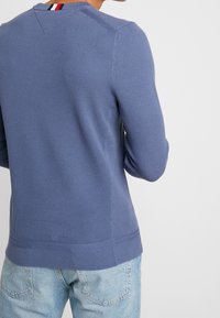 Blå stickad tröja med ett texturerat mönster, med en ribbad nederkant och krage, accentuerad med ett rödvitt rand bak.