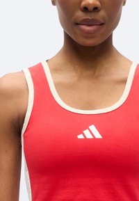 Κόκκινο αθλητικό αμάνικο με στρογγυλή λαιμόκοψη, κρεμ κορδόνι και λευκό λογότυπο στο στήθος. Το ύφασμα φαίνεται ομαλό με εφαρμοστό κόψιμο.