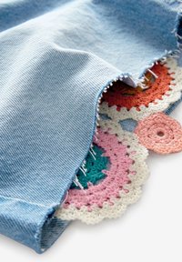 Tissu en denim bleu clair avec un bord effiloché révélant des patches circulaires colorés au crochet en dessous sur fond blanc.