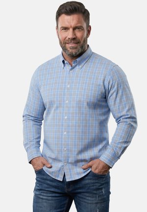 Mand med skæg iført en blå ternet skjorte med knapper og blå jeans, stående med hænderne i lommerne mod en ensfarvet baggrund.