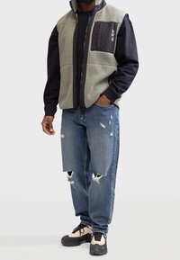Man in een lichtgrijze fleece vest over een zwart langärmige shirt, gescheurde blauwe jeans en beige en zwarte sneakers, staande met één hand in zijn zak.