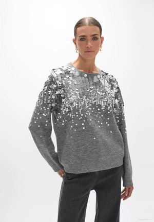 Mujer con suéter gris cubierto con grandes lentejuelas plateadas en la mitad superior, combinado con pantalones gris oscuro, de pie frente a un fondo liso.