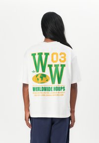 Fehér póló zöld és sárga felirattal: "WOW 03 WORLDWIDE HOOPS" földgömb grafikával és városnevekkel, rövid ujjú és laza fazonnal.