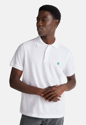 Weißes Poloshirt mit kurzen Ärmeln, weicher Textur und kleinem grünen Logo auf der linken Brust. Getragen mit grauen Jeans.