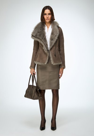 Veste en daim marron avec col en fourrure grise, chemisier blanc, jupe olive, collants noirs et talons noirs, tenant un sac à main marron texturé.