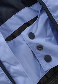 Indvendig på en lyseblå stofjakke med sort foring. Funktionen inkluderer en syet lomme, to sorte trykknapper og en lynlåslukning.