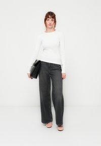 White long-sleeve top, black wide-leg jeans, holding a black clutch bag, with light-colored shoes, against a plain white background.