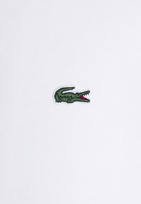 Petit logo de crocodile vert avec une bouche rouge ouverte brodée sur un tissu blanc.