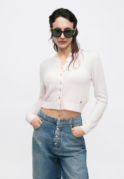 Világos rózsaszín, gombos, rövidített kardigán, bordás mandzsettákkal és apró logóval. Magas derekú, kék farmernadrág fölött viselve, amely gombos elülső résszel rendelkezik.