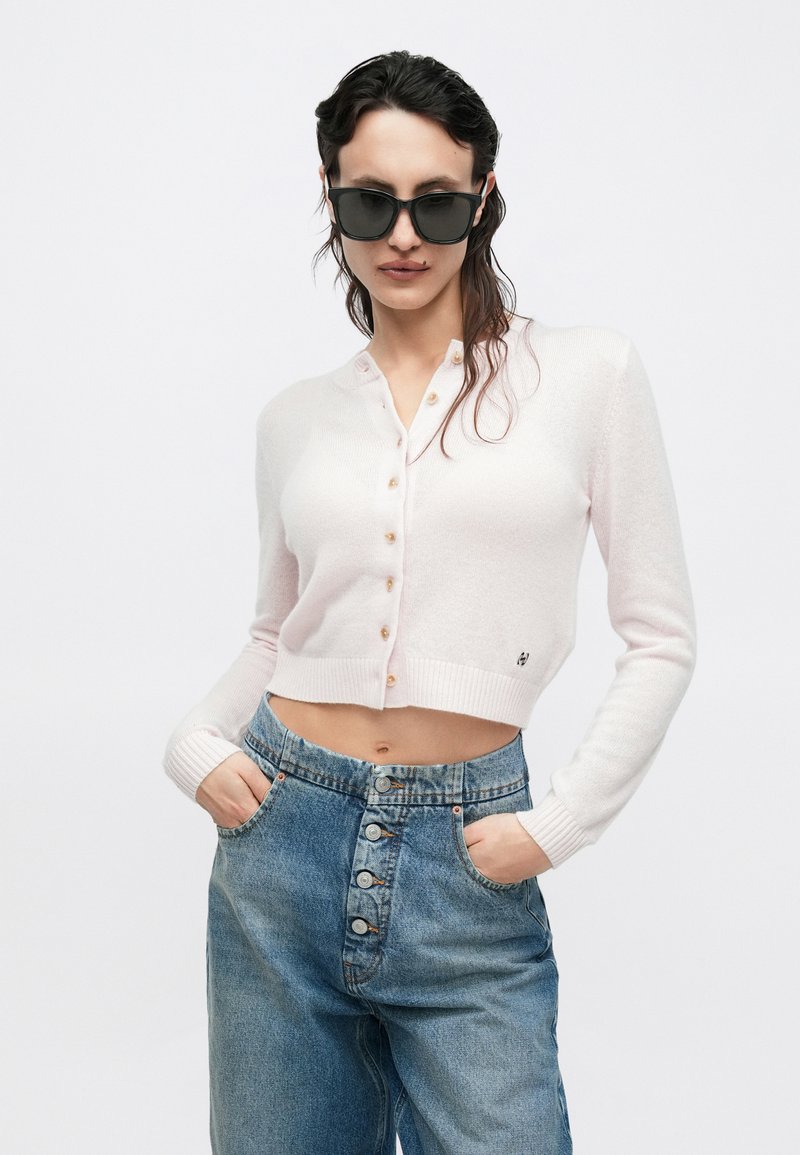 Hellrosa kurz geschnittener Cardigan mit Knöpfen, gerippten Bündchen und kleinem Logodetail. Getragen über blauem, hoch tailliertem Denim-Hose mit Knopfleiste.