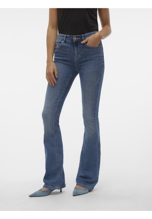 Kobieta w niebieskich jeansach bootcut, czarnej bluzce i niebieskich szpilkach w szpic, stoi z jedną ręką w kieszeni na jasnym tle.