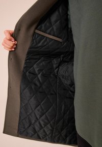 Abrigo de tela exterior gris con un interior negro acolchado. Cuenta con un bolsillo elegante y una textura suave. Sin botones ni cierres visibles.