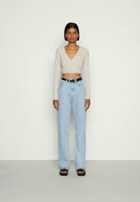Mulher jovem com cabelo curto, usando um suéter cropped em V, bege, jeans de cintura alta em azul claro, cinto preto e sandálias pretas, em pé.