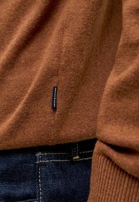 Brązowy sweter z dzianiny o miękkiej fakturze, z małą czarną metką przyszytą z boku, w połączeniu z ciemnoniebieskimi jeansami z denimu.