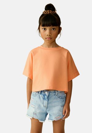 T-shirt court à manches courtes de couleur orange pastel doux, associé à un short en denim déchiré bleu clair avec des ourlets effilochés et des poches visibles.