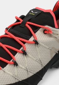 Schnürsportschuh mit einem schwarzen und beigen strukturierten Obermaterial, auffälligen roten Schnürsenkeln, silbernem Zubehör und einer Gummisohle für Trittsicherheit.