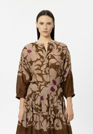 Woman with long dark hair wears a brown and beige dress with purple floral patterns, standing against a plain white background.