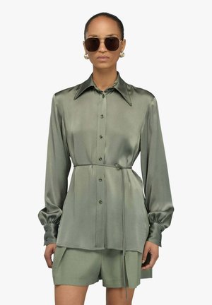 Donna che indossa una camicetta lucida verde oliva con bottoni e cintura, pantaloncini coordinati, grandi orecchini dorati e occhiali da sole grandi e scuri.