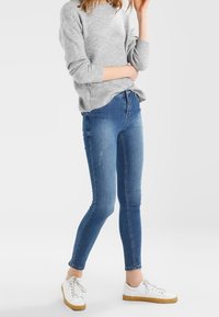 Jean skinny en denim bleu à lavage moyen, avec une taille haute, des détails subtils d'usure, et une texture lisse, associé à des baskets blanches.