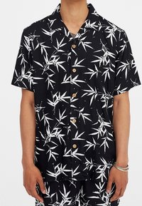 Camisa de manga corta en tela negra con un estampado de hojas de bambú en blanco, botones de madera y un corte relajado. Textura suave y diseño casual.
