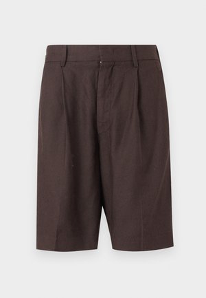 Mężczyzna stojący z rękami w kieszeniach, ubrany w paskowaną koszulę z krótkim rękawem, białą podkoszulkę, ciemnobrązowe krótkie spodenki i ciemne sandały.