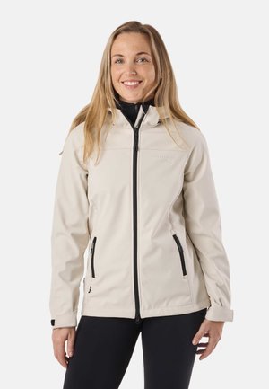 Young woman smiling, wearing light beige zip-up jacket with black zipper and pockets, standing against plain white background.