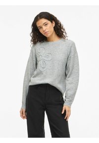 Grijze gebreide trui met een bloemmotief in lichtere grijstinten, ronde hals en lange mouwen, gecombineerd met zwarte broek. Getextureerde stof.