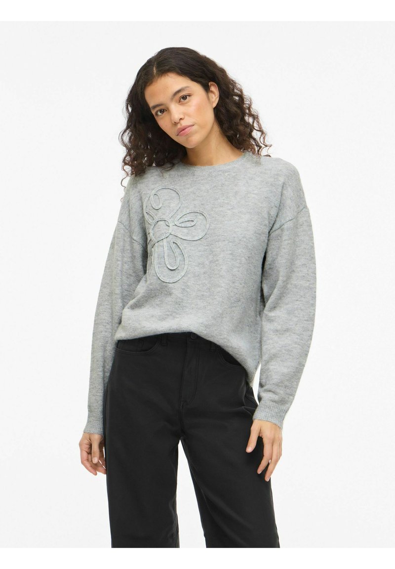 Grijze gebreide trui met een bloemmotief in lichtere grijstinten, ronde hals en lange mouwen, gecombineerd met zwarte broek. Getextureerde stof.