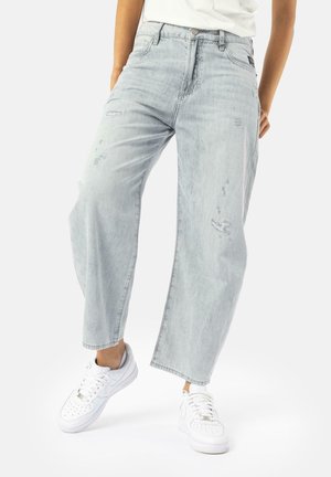 Henkilö pukeutunut vaalean pestyihin, väljästi istuviin kulutettuihin farkkuihin ja valkoisiin tennareihin, seisoo rentona asennossa yksivärisen valkoisen taustan edessä.