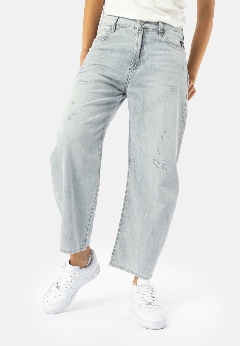 Personne portant un jean clair délavé, ample et usé ainsi que des baskets blanches, debout dans une posture détendue devant un fond blanc uni.