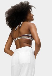 Woman with voluminous curly hair wearing a patterned halter bikini top and loose white pants, standing with hands in pockets.