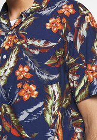 Lyhythihainen paita tummansinisestä kankaasta, jossa on kirkkaan oransseja kukkia ja vihreitä lehtiä. Mukana kaulus ja valkoiset napit.