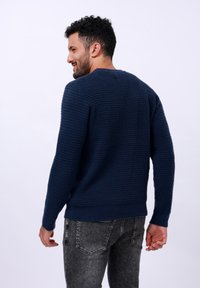 Pull en mailles bleu marine texturé avec un col rond et des poignets côtelés. Présente un ourlet qui se situe à la taille, associé à un jean gris.