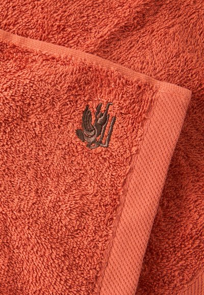 Gros plan d'une serviette en tissu éponge orange corail avec une bordure texturée et un petit motif de main brodé foncé.
