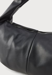 Musta keinonahkainen olkalaukku, jossa on hopeinen vetoketju ja rypytetty muotoilu valkoisella taustalla.