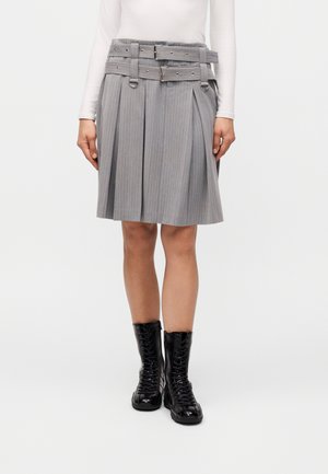 Vrouw draagt een grijze geplooide pinstripe rok met dubbele riemdetail en zwarte lakleren veterlaarzen, staand tegen een witte achtergrond.