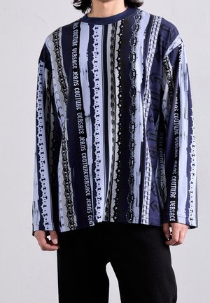 Mężczyzna ubrany w koszulę z długim rękawem w pionowe paski z motywem łańcuchów oraz napisem "Versace Jeans Couture" w niebieskich, czarnych i białych tonacjach.