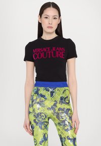 Camiseta negra de manga corta con "VERSACE JEANS COUTURE" en texto fucsia en negrita, combinada con pantalones amarillos brillantes con lentejuelas y acentos azules.