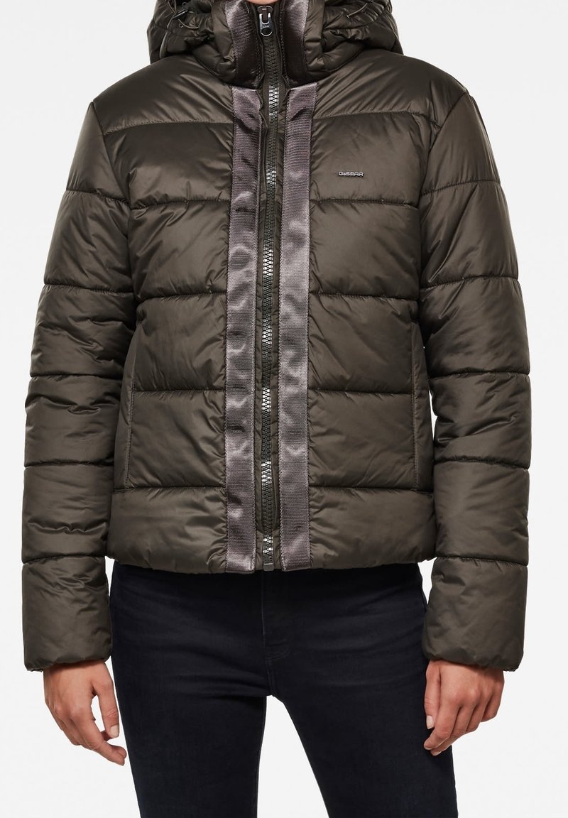 Personne portant une doudoune brillante vert foncé avec capuche et bandes grises verticales le long de la fermeture éclair devant, associée à un pantalon noir.
