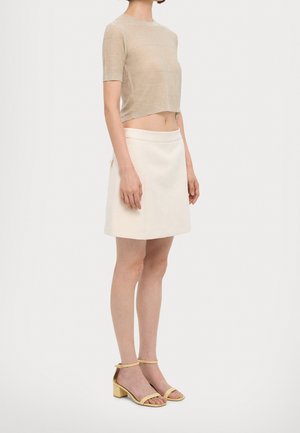 Femme portant un haut en maille beige à manches courtes, une jupe couleur crème à hauteur des genoux et des sandales jaunes à talons blocs ouvertes, debout de profil.