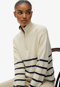 Jersey crema con cuello alto y medio cierre, con rayas horizontales en azul marino y un patrón de tejido texturizado. Detalle sutil del logo en el pecho.