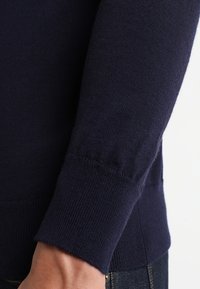Granatowy sweter o gładkiej fakturze, dopasowanych mankietach i prostym, eleganckim wzorze. Tkanina wydaje się miękka i dziana.
