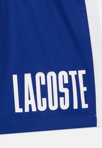 Kék sport rövidnadrág légáteresztő hálós anyagból. Fehér oldalsó díszítéssel és nagy, vastag betűs "LACOSTE" logóval a szegélyen.
