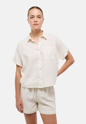 Femme portant une chemise beige clair à manches courtes boutonnée et un short assorti à fines rayures verticales, debout avec une main sur la hanche.