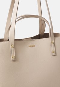 Bežová kožená taška přes rameno s texturovaným povrchem, dvojitými popruhy a zlatými kovovými prvky. Název značky je vyražený na přední straně.