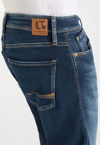 Jeans de mezclilla azul oscuro con un acabado texturizado, que presentan un parche de logo de cuero en la cintura y detalles de costura visibles.